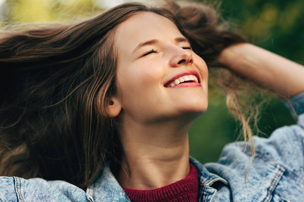 Retrato de cerca de una hermosa joven sonriendo ampliamente con una sonrisa dentada saludable, con los ojos cerrados, con chaqueta de mezclilla, posando sobre el fondo de la naturaleza en el parque. Personas, viajes, estilo de vida
 - Foto, Imagen