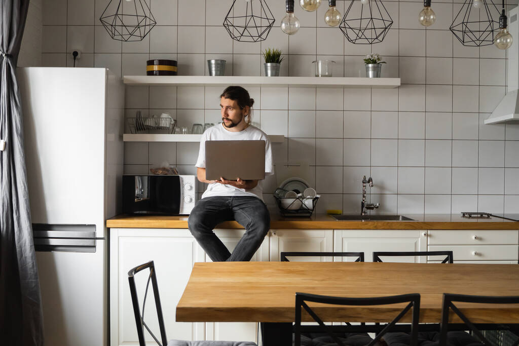 Freelancer working from home sitting on a kitchen worktop and using laptop. Bearded man working with a laptop and reading news. Handsome successful self entrepreneur working at his modern home. - Photo, Image