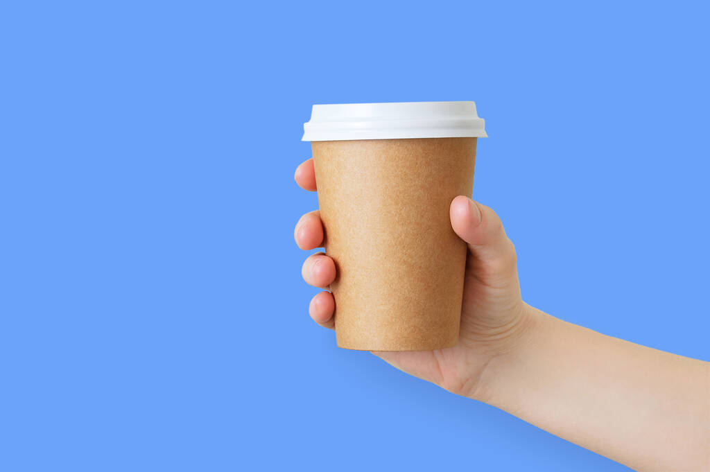 Paper brown cup of coffee in the hand of a caucasian girl. Blue background. - Photo, Image