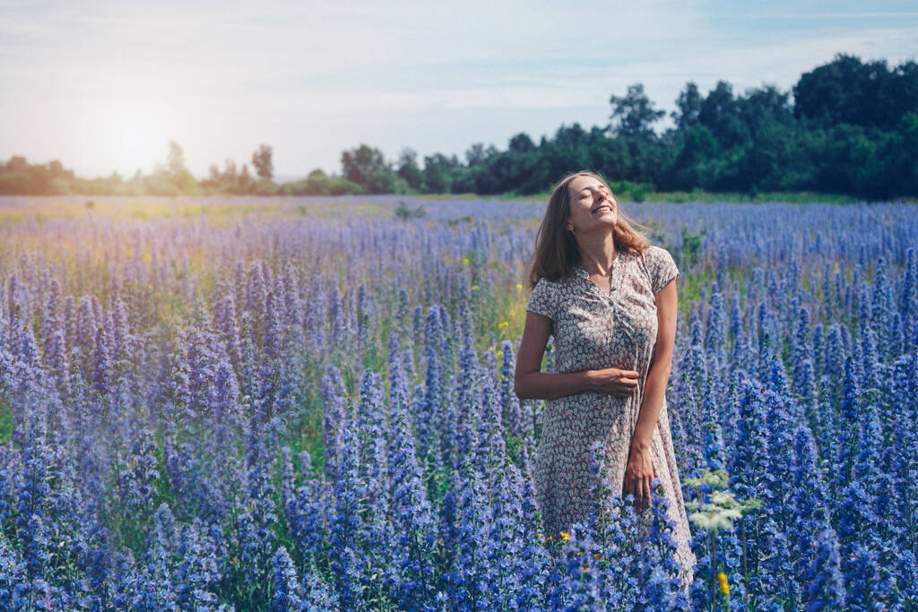 dziewczyna w luźnej sukience z rozpuszczonymi włosami przechodzi przez łąkę kwiatów. Kobieta cieszy się życiem wśród fioletowych kwiatów. Koncepcja lata, ciepła, wolności i zapachu wiosny - Zdjęcie, obraz