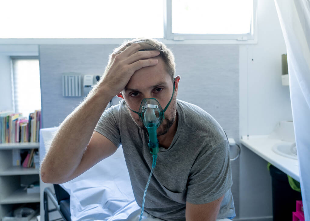 Młody atrakcyjny mężczyzna z maską tlenową wyglądający smutno i zmartwiony w szpitalnym łóżku w sypialni kliniki w diagnostyce raka płuc, paleniu i chorobach układu oddechowego oraz kampanii reklamowej antytytoniowej. - Zdjęcie, obraz