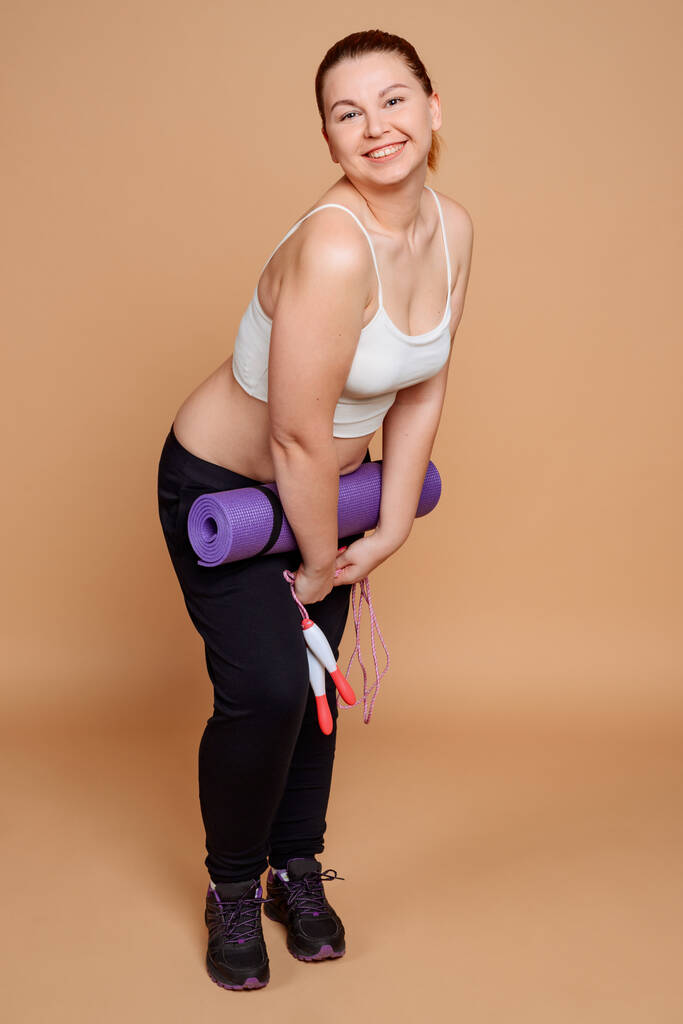 Plus size vrouw poseren met fitnessapparatuur en heldere glimlach op beige - Foto, afbeelding