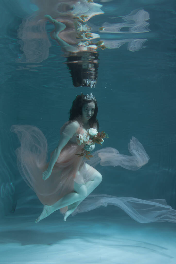 A girl with long dark hair swims underwater in a pink dress and with a crown on her head, like an underwater queen. Fairy tale suitable for advertising - Photo, Image