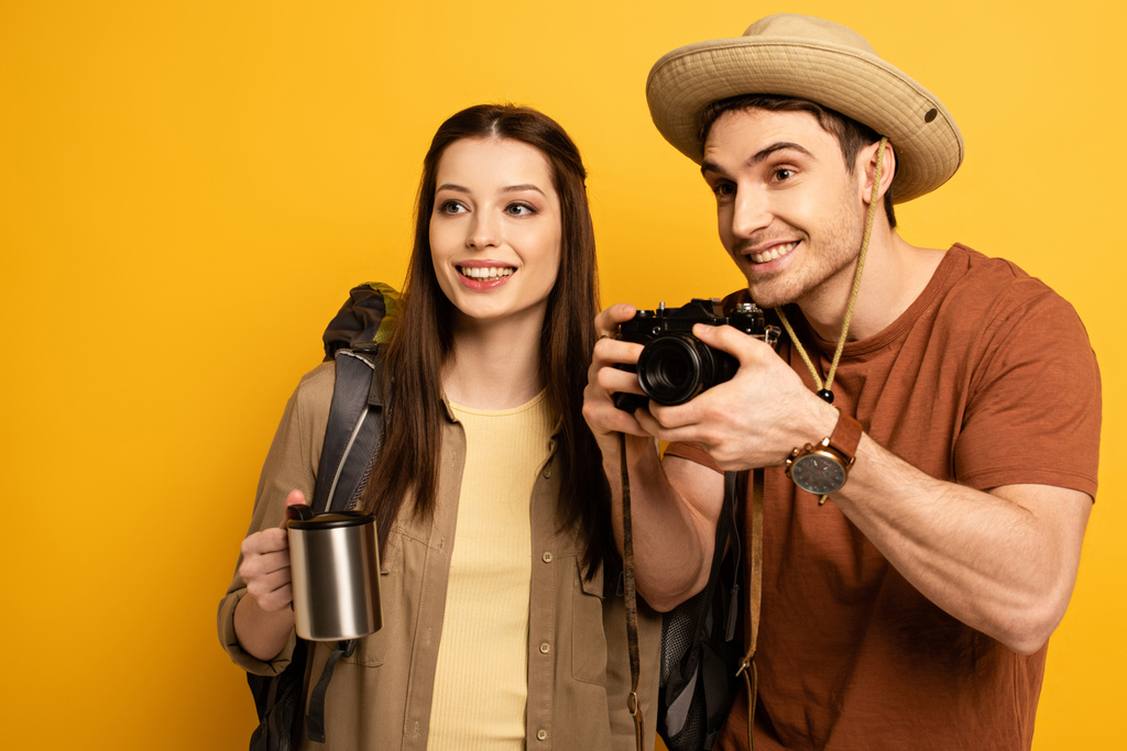 pareja de turistas sonrientes con mochilas con cámara fotográfica y café, aislados en amarillo
 - Foto, Imagen
