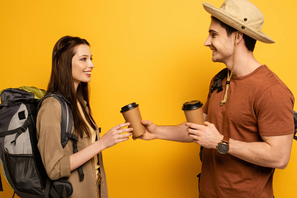 couple of happy travelers with backpacks holding coffee to go on yellow - Photo, Image