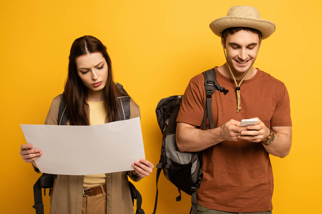 Ein paar glückliche Touristen mit Rucksäcken blicken auf Karte und Smartphone auf gelb - Foto, Bild