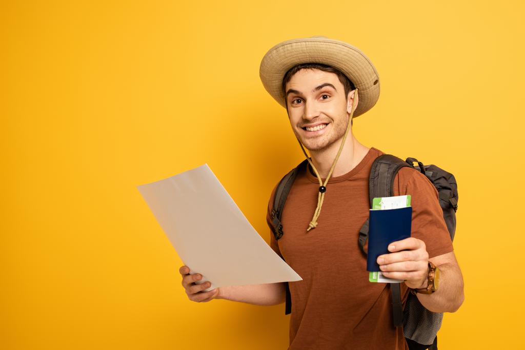 χαμογελαστός τουρίστας στο καπέλο με σακίδιο που κατέχουν διαβατήριο, εισιτήριο και χάρτη σε κίτρινο  - Φωτογραφία, εικόνα