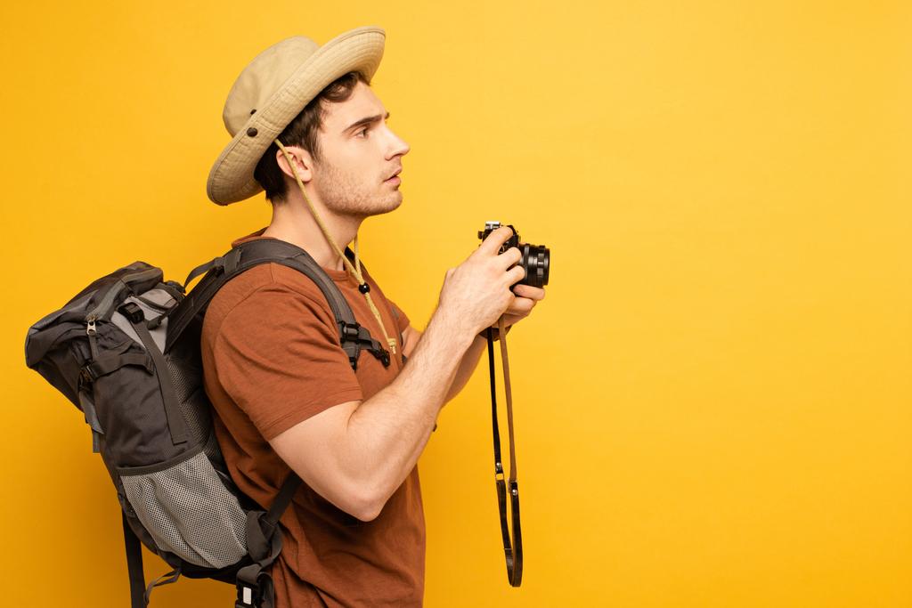 όμορφος ταξιδιώτης σε καπέλο με σακίδιο κρατώντας φωτογραφική μηχανή φωτογραφία στο κίτρινο - Φωτογραφία, εικόνα