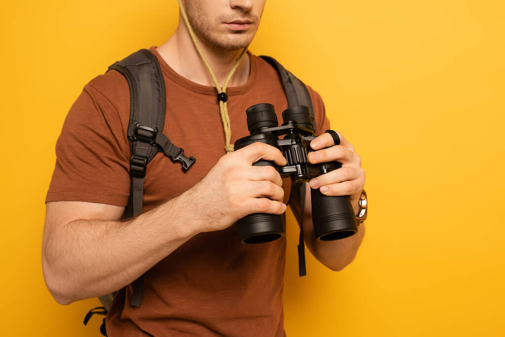 bijgesneden weergave van reiziger met rugzak met verrekijker op geel - Foto, afbeelding