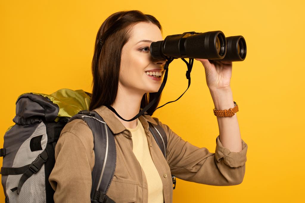 beautiful female traveler with backpack looking through binoculars, isolated on yellow  - Photo, Image