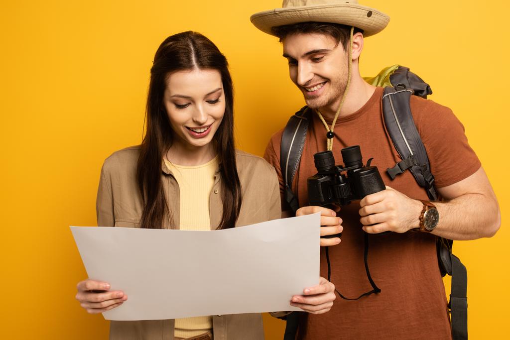 ζευγάρι χαμογελαστοί ταξιδιώτες με σακίδιο πλάτης και κιάλια κοιτάζοντας το χάρτη σε κίτρινο - Φωτογραφία, εικόνα