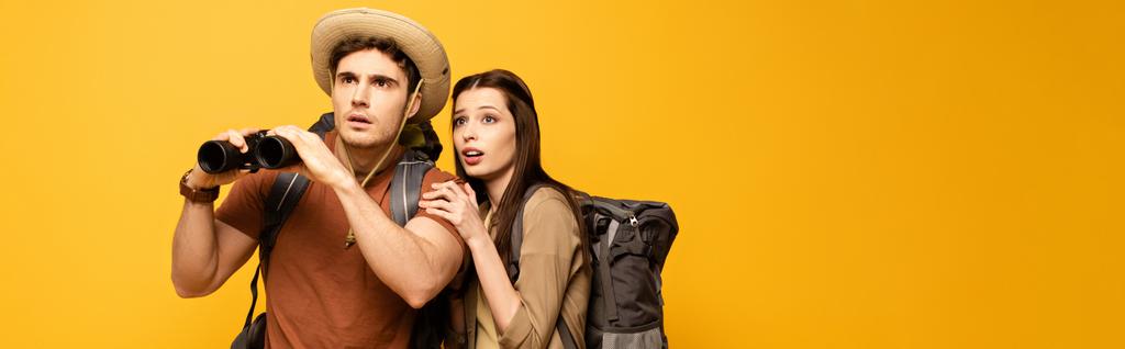 panoramiczne ujęcie dwóch zszokowanych turystów z plecakami i lornetką, odizolowanych na żółto - Zdjęcie, obraz