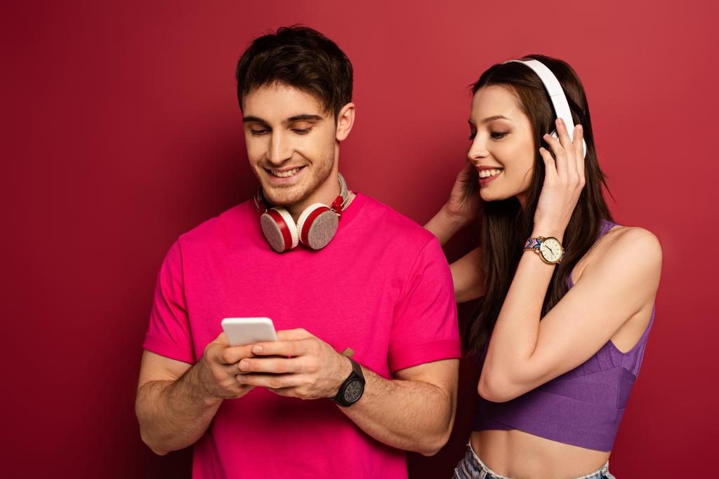 hermosa pareja positiva con auriculares usando teléfono inteligente en rojo
 - Foto, Imagen