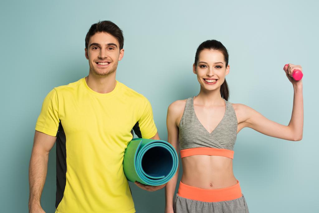 χαρούμενο αθλητικό ζευγάρι κρατώντας στρώμα γυμναστικής και αλτήρα σε μπλε - Φωτογραφία, εικόνα