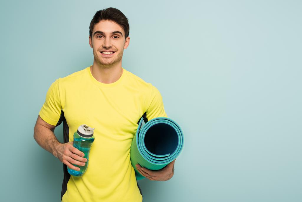 fröhlicher muskulöser Sportler im gelben T-Shirt mit Fitnessmatte und Sportflasche mit Wasser auf blau - Foto, Bild