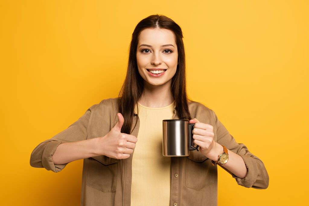 χαρούμενο θηλυκό ταξιδιώτη κρατώντας φλιτζάνι καφέ και δείχνοντας τον αντίχειρα επάνω στο κίτρινο  - Φωτογραφία, εικόνα