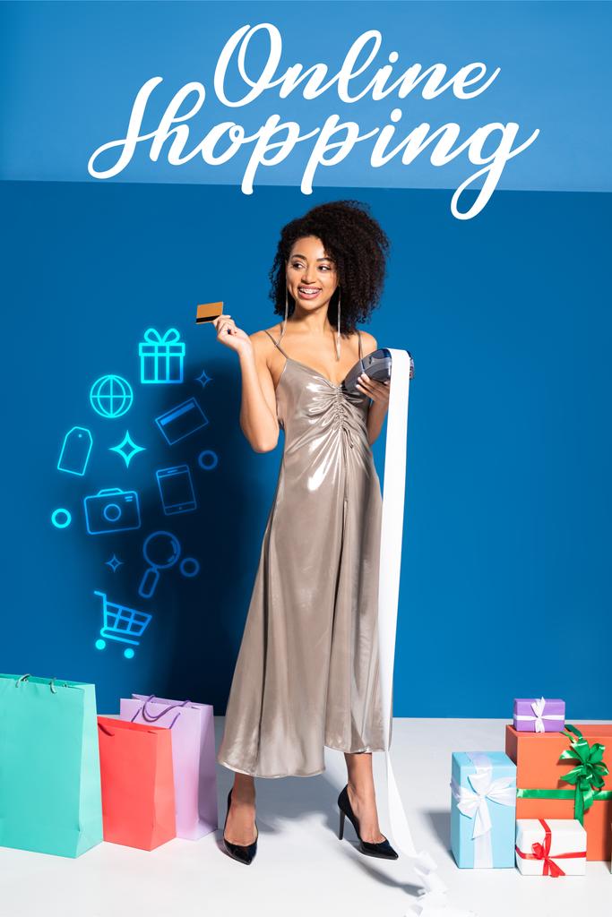 ευτυχισμένη όμορφη Αφροαμερικανή γυναίκα σε ασημί φόρεμα κρατώντας τερματικό πληρωμής με επιταγή και πιστωτική κάρτα κοντά σε δώρα και τσάντες ψώνια σε μπλε φόντο, σε απευθείας σύνδεση απεικόνιση ψώνια - Φωτογραφία, εικόνα