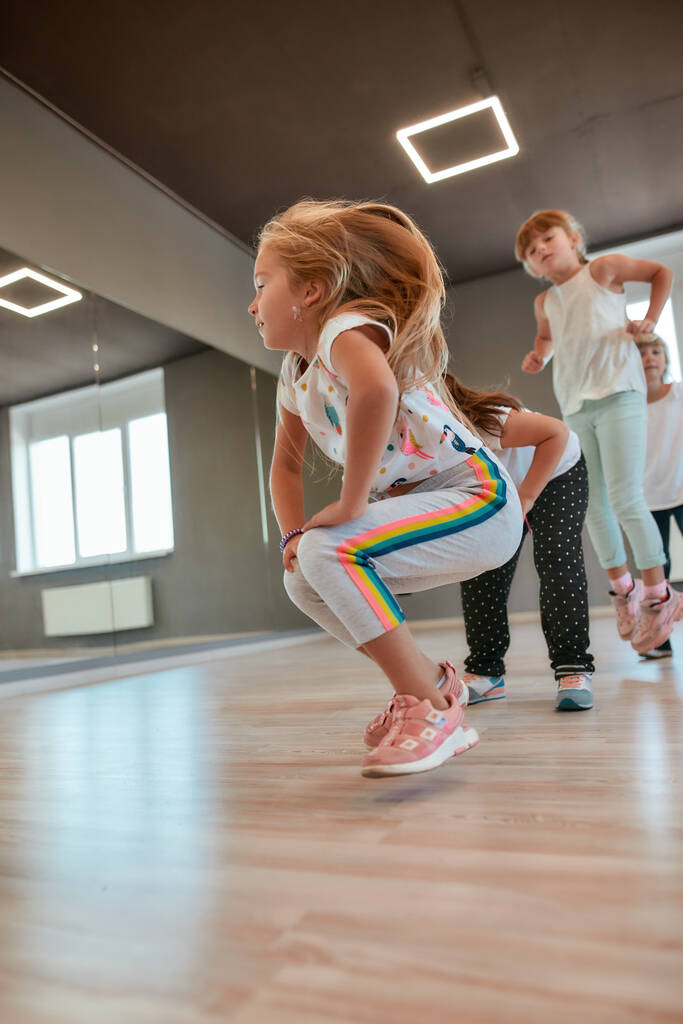 Динамичная разминка. Маленькие активные мальчики и девочки в повседневной одежде разогреваются в танцевальной студии. Дети делают спортивные упражнения
 - Фото, изображение