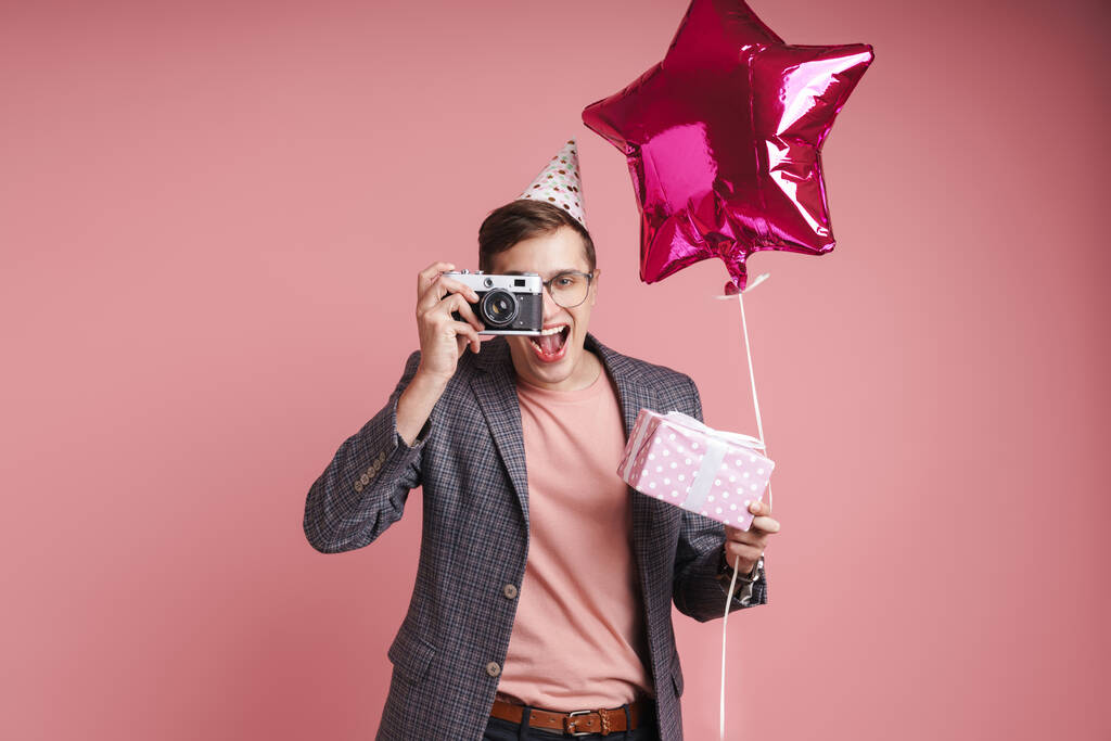 Portrét mladého muže s narozeninovým kloboukem, který slaví izolaci nad růžovým pozadím, drží dárkovou krabici a balónek, fotí se fotoaparátem - Fotografie, Obrázek