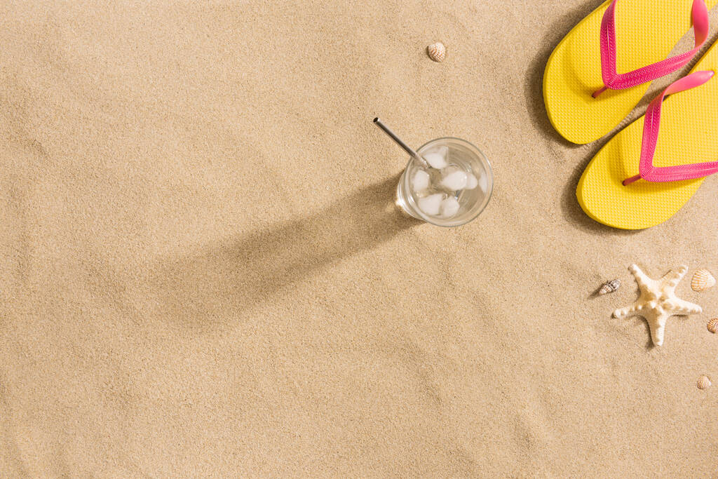 夏のファッション、砂の背景に夏服。黄色のフリップフロップ、水と貝殻のガラス。フラットレイアウト、トップビュー。影の強い光 - 写真・画像