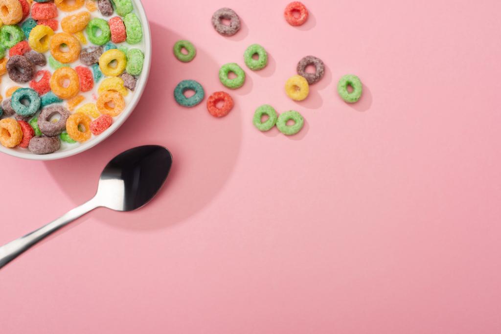 Draufsicht auf helle bunte Frühstückscerealien mit Milch in Schüssel in der Nähe von Löffel auf rosa Hintergrund - Foto, Bild
