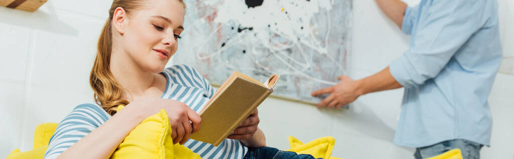 Foco seletivo da mulher ler livro perto namorado segurando pintura na sala de estar, tiro panorâmico
 - Foto, Imagem
