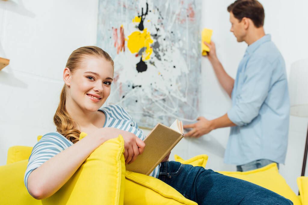 Selectieve focus van de vrouw glimlachen op de camera tijdens het lezen van boek in de buurt vriendje met doek schoonmaken schilderen thuis - Foto, afbeelding