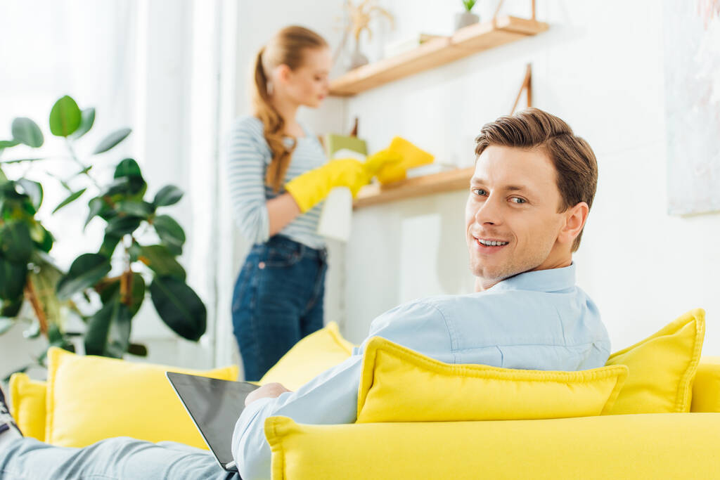 Szelektív fókusz férfi laptop mosolyog a kamera, miközben ül a kanapén közelében barátnője takarítás polc a nappaliban - Fotó, kép