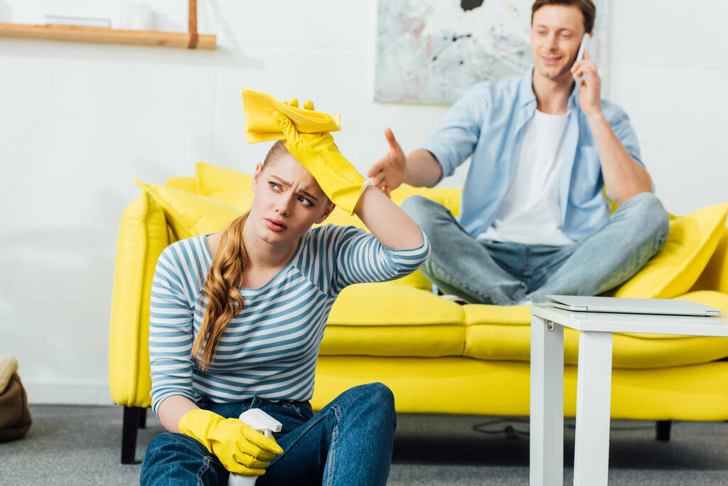 Enfoque selectivo de la mujer cansada con suministros de limpieza sentado en el suelo cerca de novio hablando en el teléfono inteligente en el sofá en casa
 - Foto, Imagen