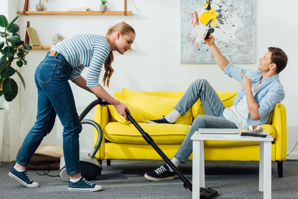 Side view of man taking selfie with smartphone on couch while girlfriend cleaning carpet with vacuum cleaner at home - Photo, Image