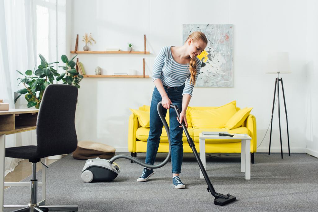 Mulher sorridente tapete de limpeza com aspirador de pó na sala de estar
 - Foto, Imagem