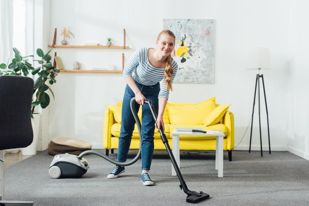 Donna attraente sorridente alla fotocamera durante la pulizia tappeto con aspirapolvere in soggiorno
 - Foto, immagini