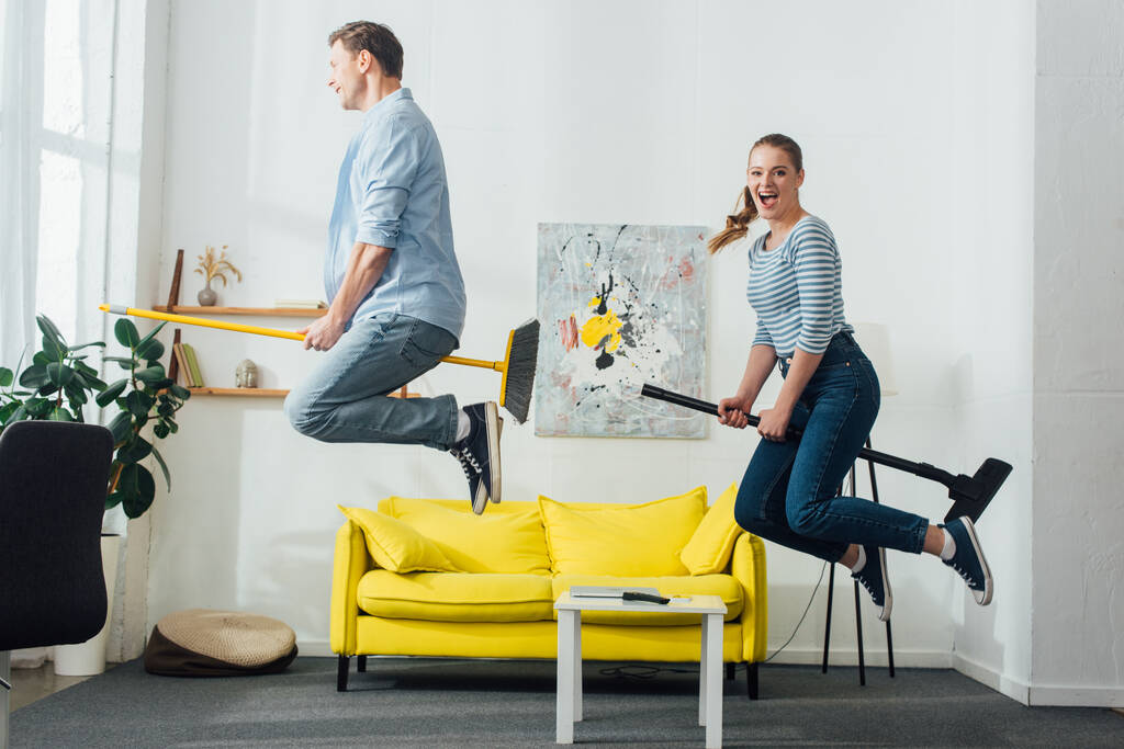 Vista lateral de casal alegre levitando em vassouras na sala de estar
 - Foto, Imagem