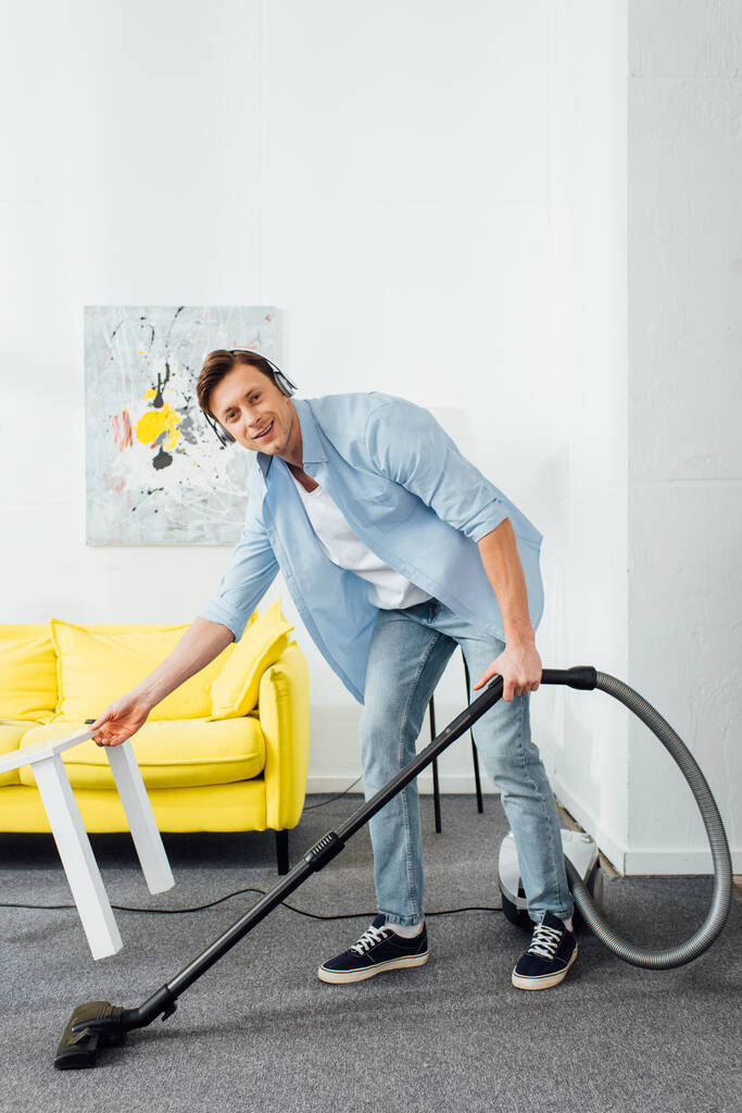 Homme souriant dans les écouteurs regardant la caméra tout en nettoyant tapis avec aspirateur dans le salon
 - Photo, image