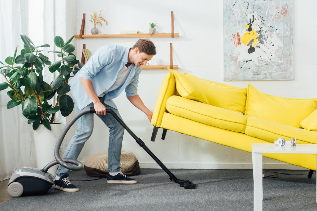 Vue latérale de l'homme soulevant le canapé tout en nettoyant le tapis avec aspirateur dans le salon
 - Photo, image
