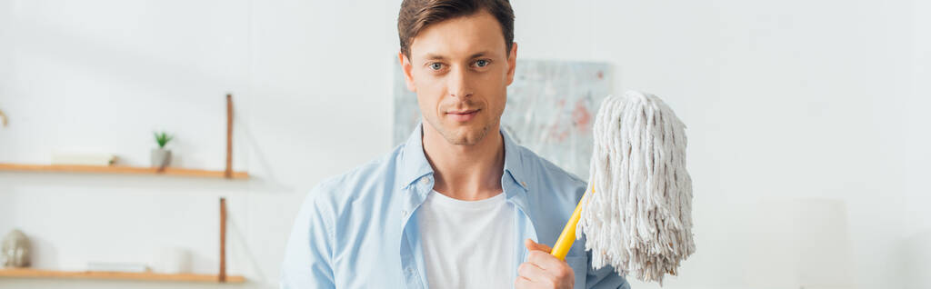 Handsome man looking at camera and holding mop in living room, panoramic shot  - Photo, Image