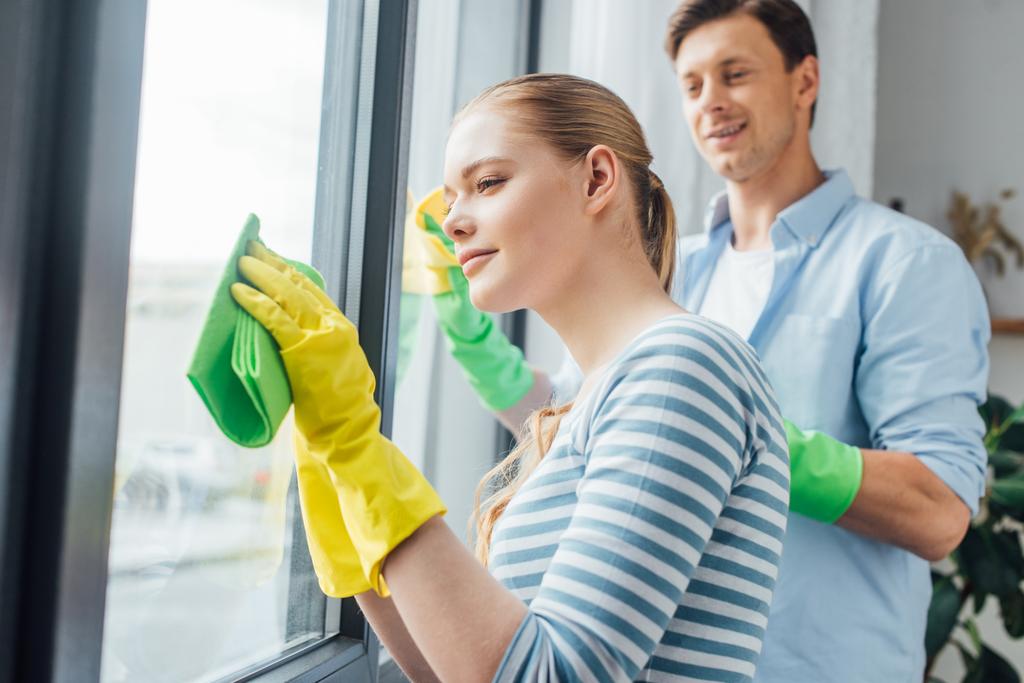 Selectieve focus van jonge vrouw schoonmaken venster in de buurt glimlachend vriendje in de woonkamer - Foto, afbeelding