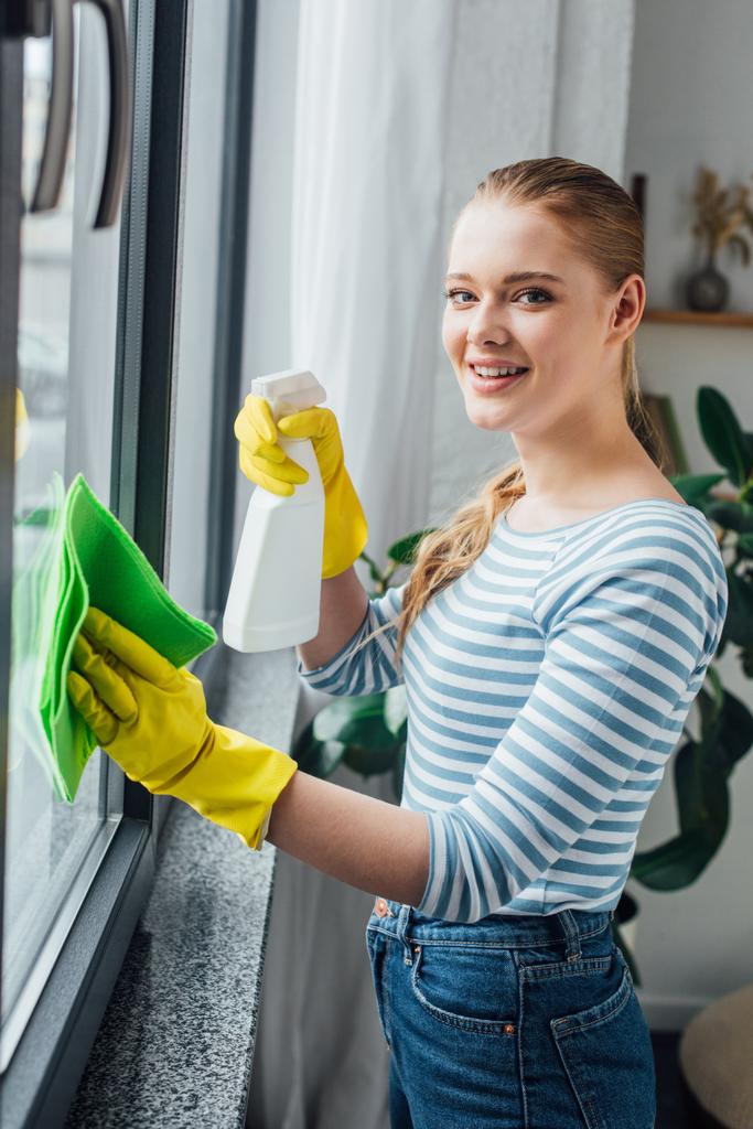 Widok z boku uśmiechnięta kobieta patrząc w kamerę podczas czyszczenia okna w domu - Zdjęcie, obraz
