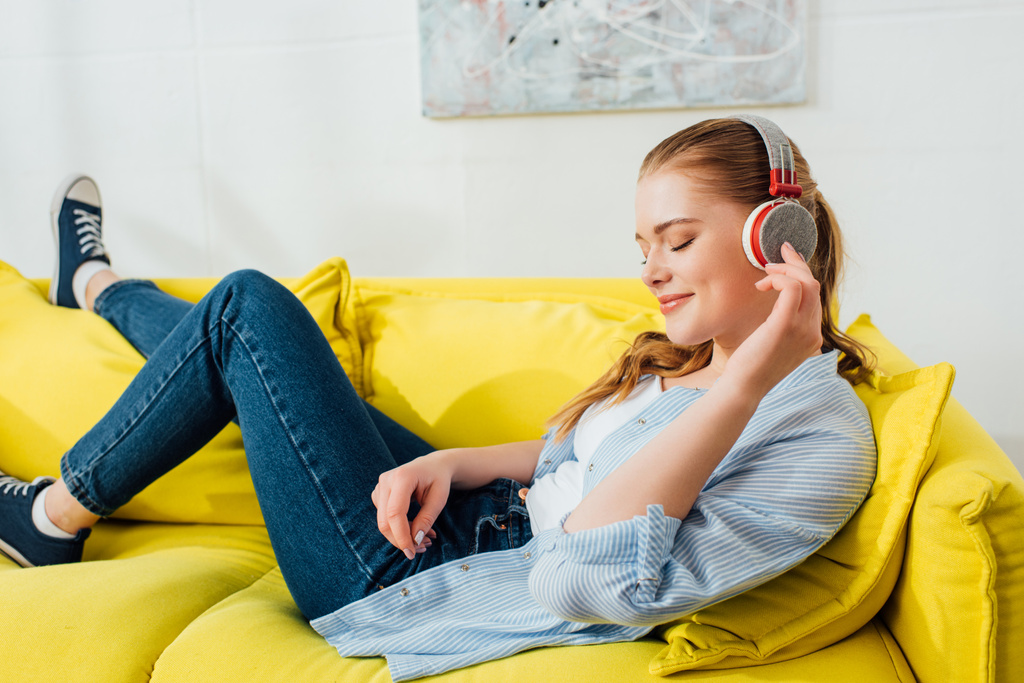Вид сбоку улыбающейся девушки в наушниках, слушающей музыку на диване
  - Фото, изображение