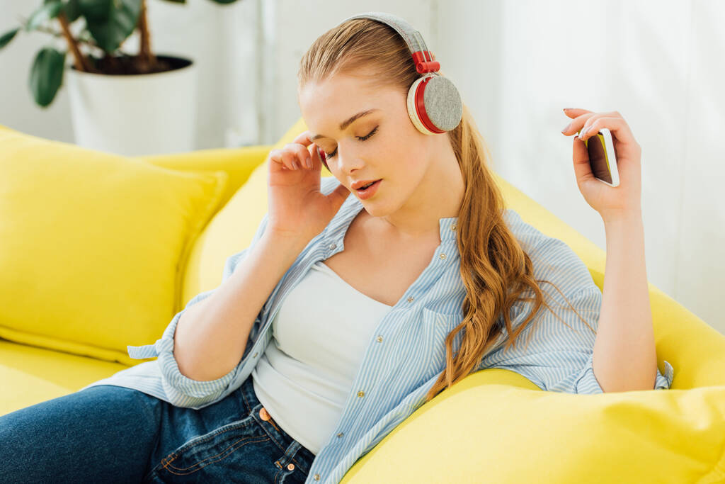 Mulher atraente com olhos fechados ouvindo música em fones de ouvido no sofá
  - Foto, Imagem