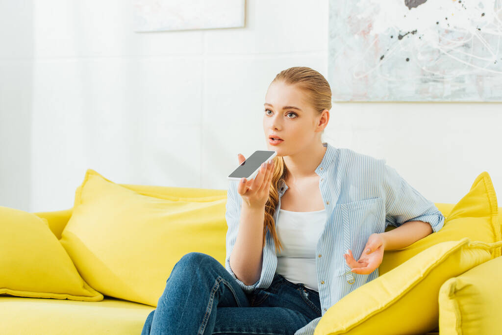 Mulher atraente usando alto-falante enquanto fala no smartphone no sofá em casa
  - Foto, Imagem