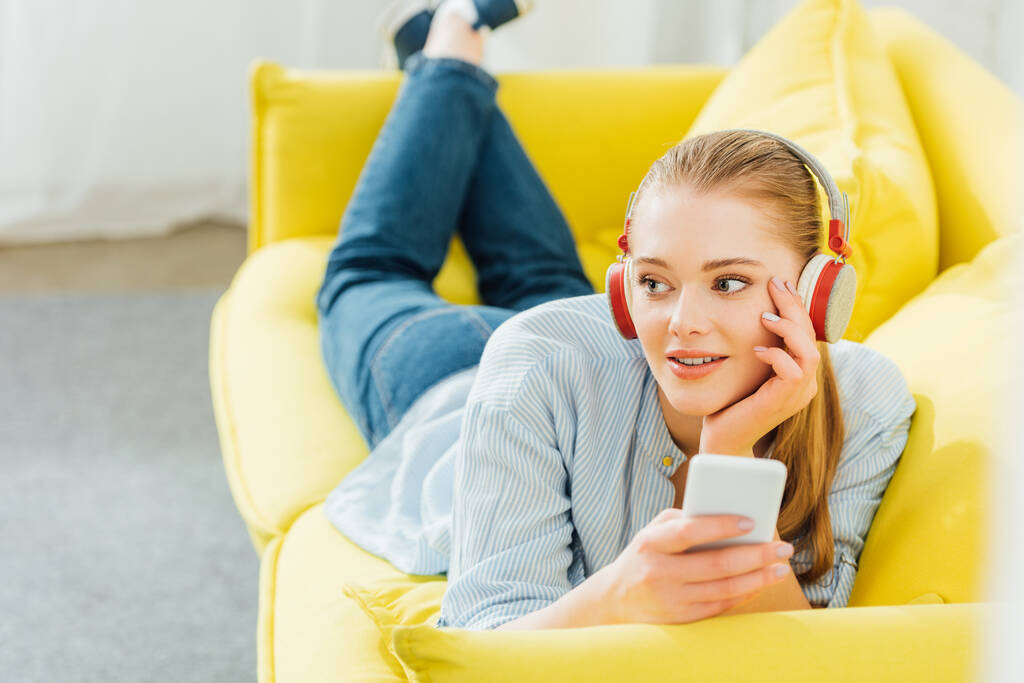Επιλεκτική εστίαση της όμορφης γυναίκας στα ακουστικά κρατώντας smartphone, ενώ βρίσκεται στον καναπέ  - Φωτογραφία, εικόνα