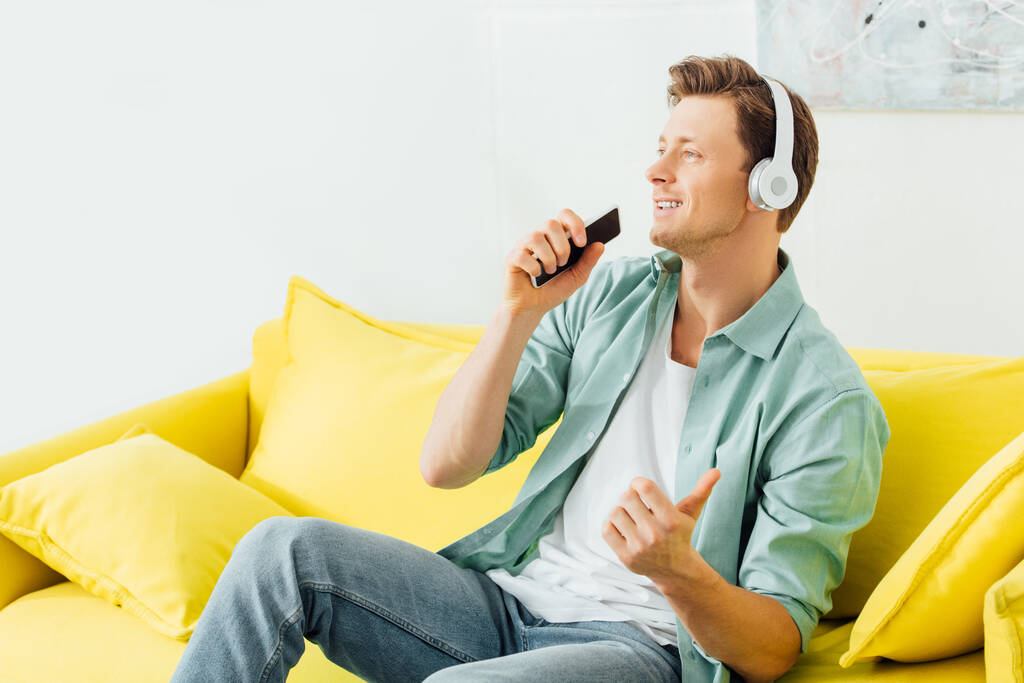 Hezký muž ve sluchátkách drží smartphone na gauči doma - Fotografie, Obrázek