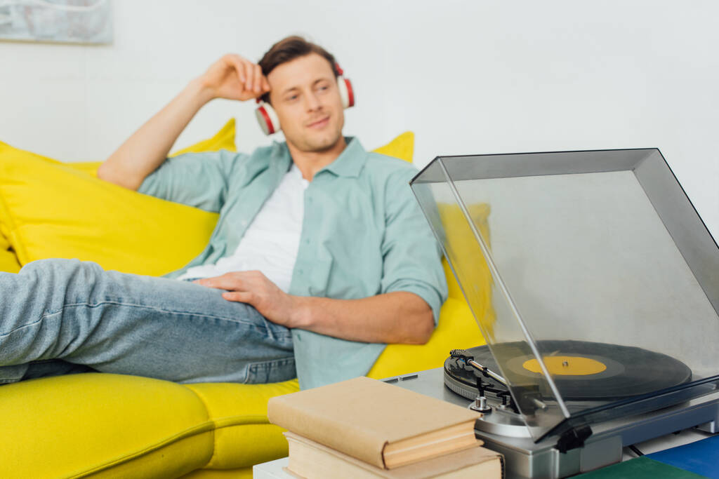 Selektiivinen painopiste levysoitin ja kirjoja sohvapöytä ja mies kuulokkeet istuu sohvalla kotona
 - Valokuva, kuva