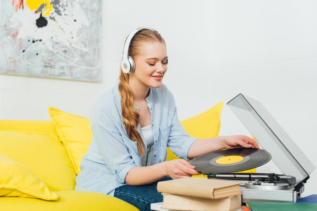 Lächelnde Frau mit Kopfhörer hält Schallplatte neben Plattenspieler und Bücher auf Couchtisch  - Foto, Bild