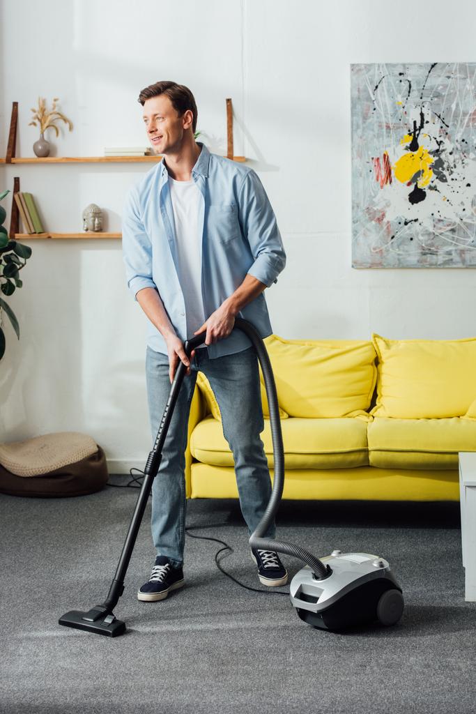 Sorrindo homem olhando para longe enquanto limpando tapete com aspirador de pó em casa
  - Foto, Imagem