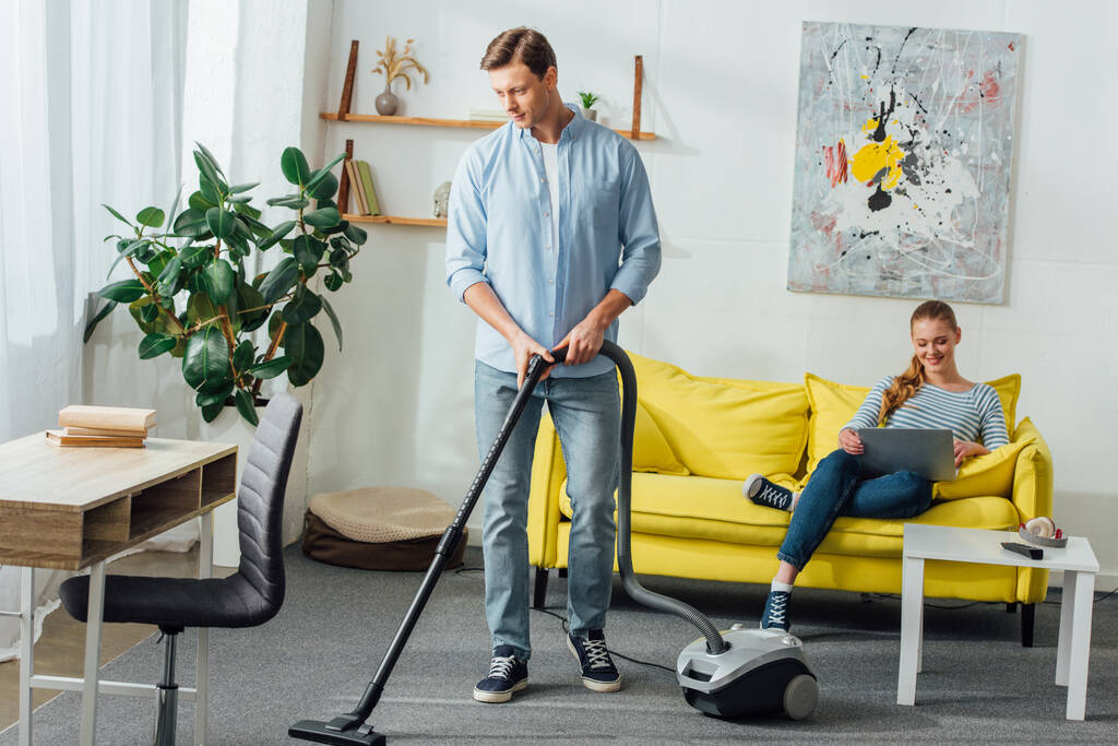 Man cleaning carpet with vacuum cleaner near smiling girl using laptop on couch at home  - Photo, Image