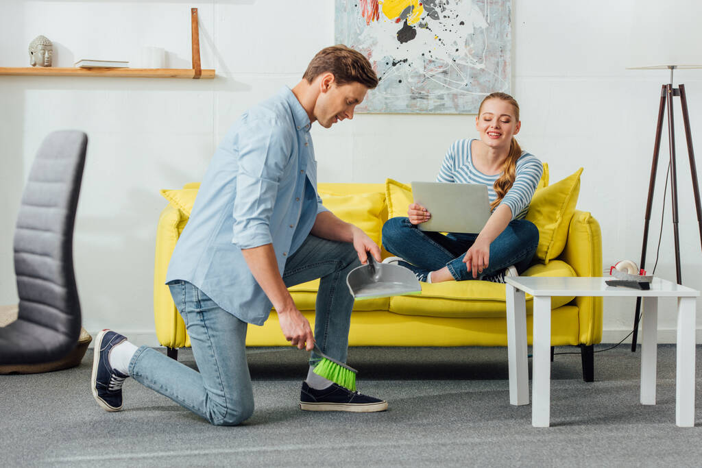 Smiling woman pointing on carpet to boyfriend with scoop and broom at home  - Photo, Image