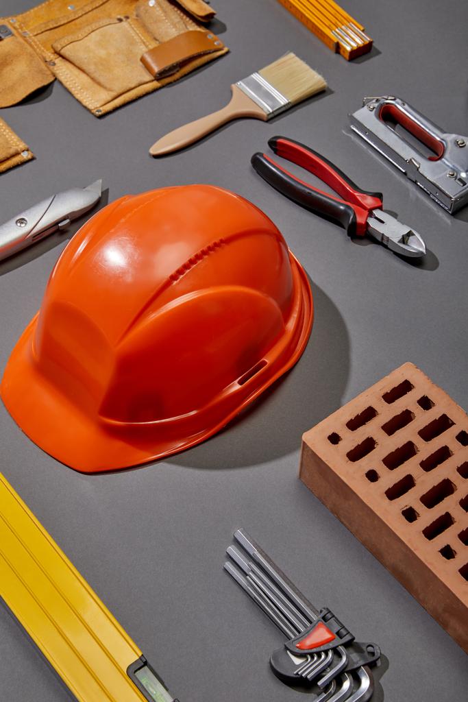 επίπεδο lay με πορτοκαλί κράνος, τούβλο, βούρτσα, τούβλο, ζώνη εργαλείων και βιομηχανικά εργαλεία σε γκρι φόντο - Φωτογραφία, εικόνα