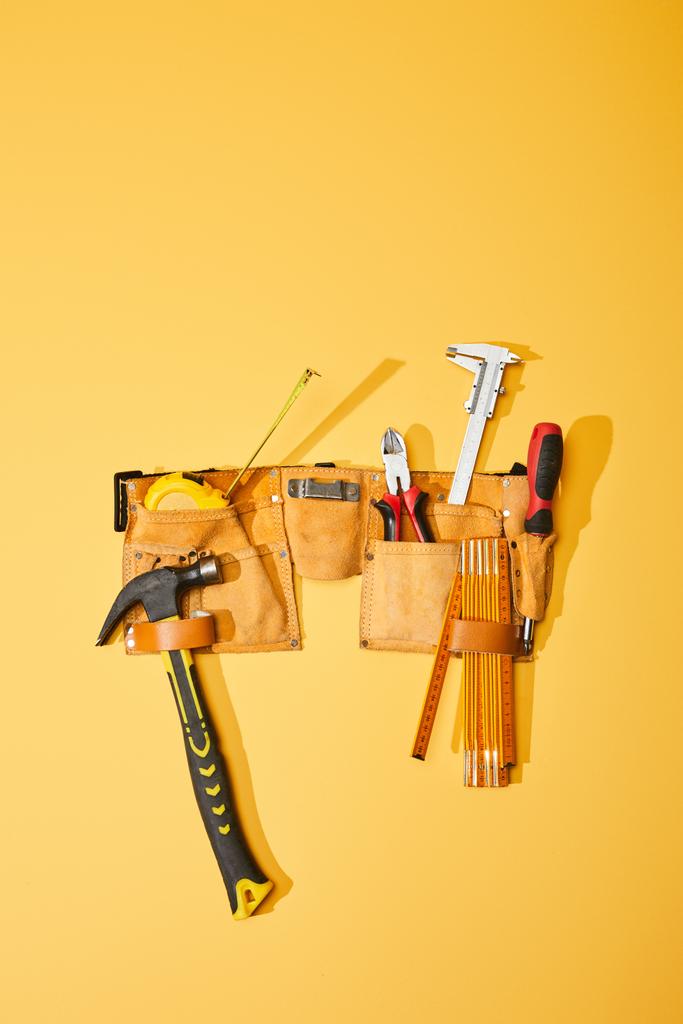 vista superior do cinto de ferramentas com martelo, fita métrica, alicate, paquímetro, chave de fenda e régua dobrável sobre fundo amarelo
 - Foto, Imagem